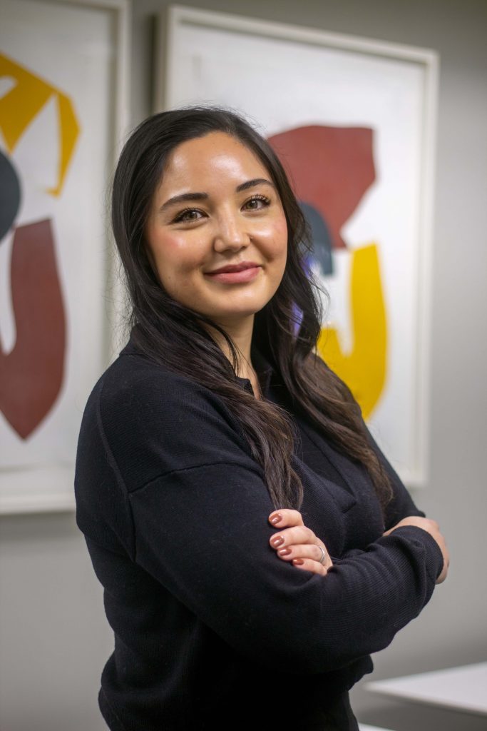 Rosanna Manaloto-Arshi stands with her arms crossed, smiling at the camera over her shoulder. She stands in front of abstract are in the CRIEC headquarters.
