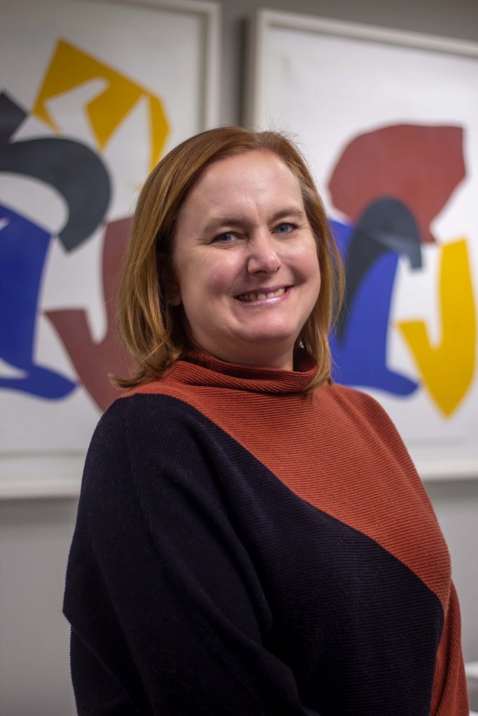 Susan Reckseidler, stands angled to the camera wearing a black and orange sweater, smiling at the camera in front of abstract art